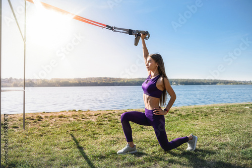 Attractive young woman doing TRX training outdoors near the lake at daytime. Healthy lifestyle.