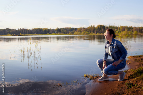Girl or woman sitting on the shore or beach of the lake in the evening at sunset. Single trip or vacation.