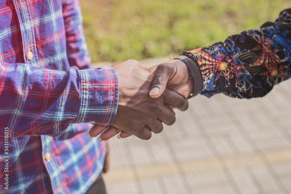 Close up view of two young african men with dark skin tone shaking hands outside in the city park in sunny weather. 