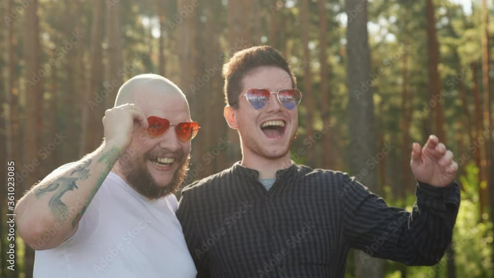 Homosexual couple smile nad laugh. Portrait of two affectionate gay men in love wearing sunglasses of heart shape smiling to camera. LGBTQI, Pride Event, LBGT Pride Month, Gay Pride Symbol. Homosexual couple smile nad laugh. Portrait of two affectionate gay men in love wearing sunglasses of heart shape smiling to camera. LGBTQI, Pride Event, LBGT Pride Month, Gay Pride Symbol.