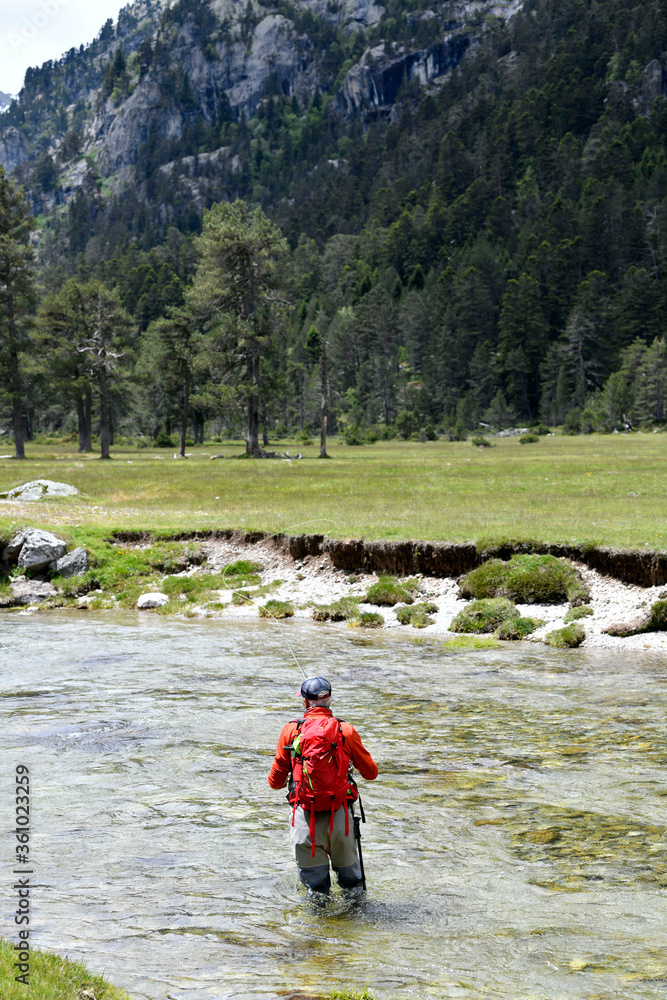 fly fisherman trout fishing with a hiking backpack and an orange jacket in the high mountains in summer
