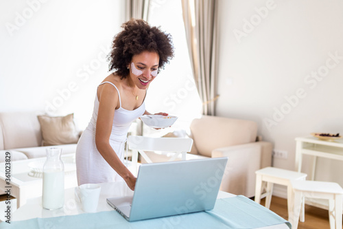 Beautiful young woman enjoys eating corn flakes for breakfast while working on her laptop in the morning.