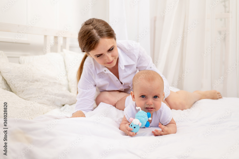 mom playing with baby 6 months lying on a white bed, leisure mom with baby, place for text mom playing with baby 6 months lying on a white bed, leisure mom with baby, place for text
