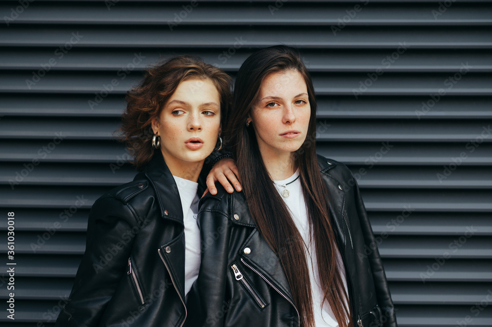 Portrait of 2 stylish models on a dark background,brunette girl looking in camera with serious face, curly girlfriend looking aside.Fashionable portrait of two girls in casual clothes on a black wall