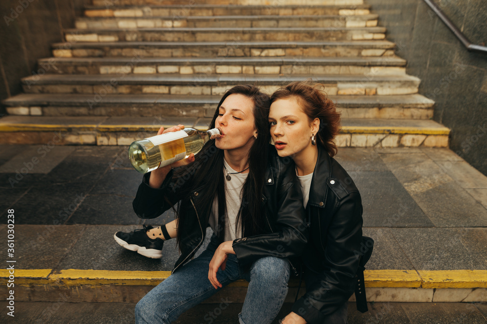 Φωτογραφία Portrait girls in leather jackets and jeans drinking wine from a bottle on the stairs in the subway and posing at camera, fashionable portrait