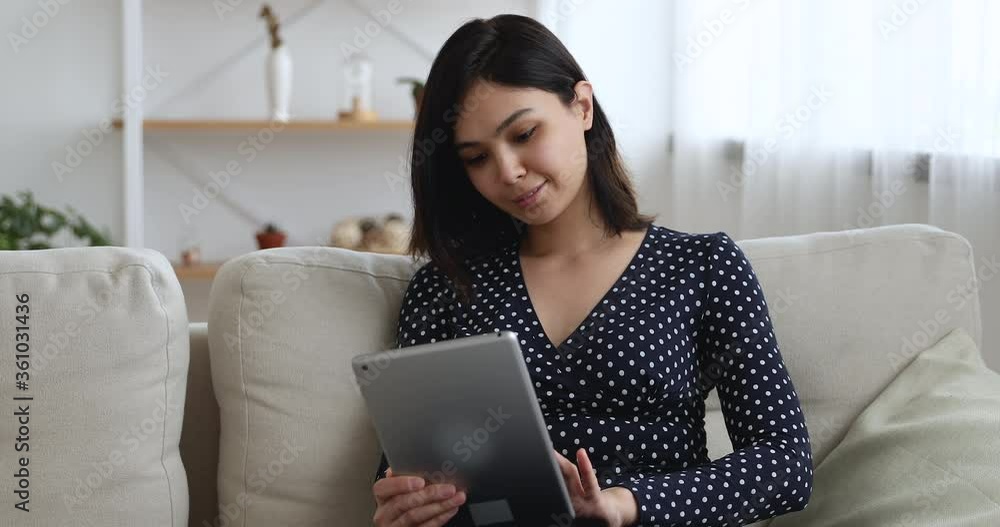 Asian woman sitting on couch in cozy living room hold tablet device smile enjoy free time chat on-line with friends, buy in e-commerce web sites. Modern tech, everyday usage easy and cool app concept Asian woman sitting on couch in cozy living room hold tablet device smile enjoy free time chat on-line with friends, buy in e-commerce web sites. Modern tech, everyday usage easy and cool app concept