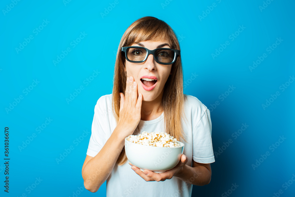 Young beautiful woman in a white T-shirt smiles with glasses for 3D and iMAX holds a plate with popcorn on a blue background. Concept of watching new movies at the cinema or at home Young beautiful woman in a white T-shirt smiles with glasses for 3D and iMAX holds a plate with popcorn on a blue background. Concept of watching new movies at the cinema or at home