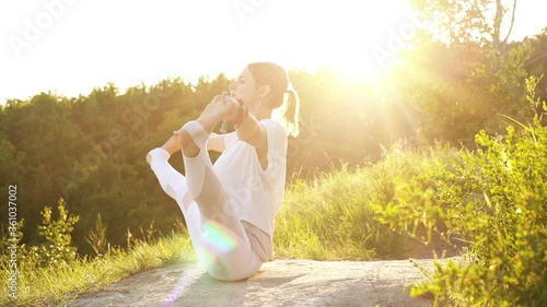 Peaceful young woman is performing yoga posture Boat with Toe Hold, outside evening on background of bright sunray. Meditative girl practicing yoga exercises in alone outdoors during sunset. photo