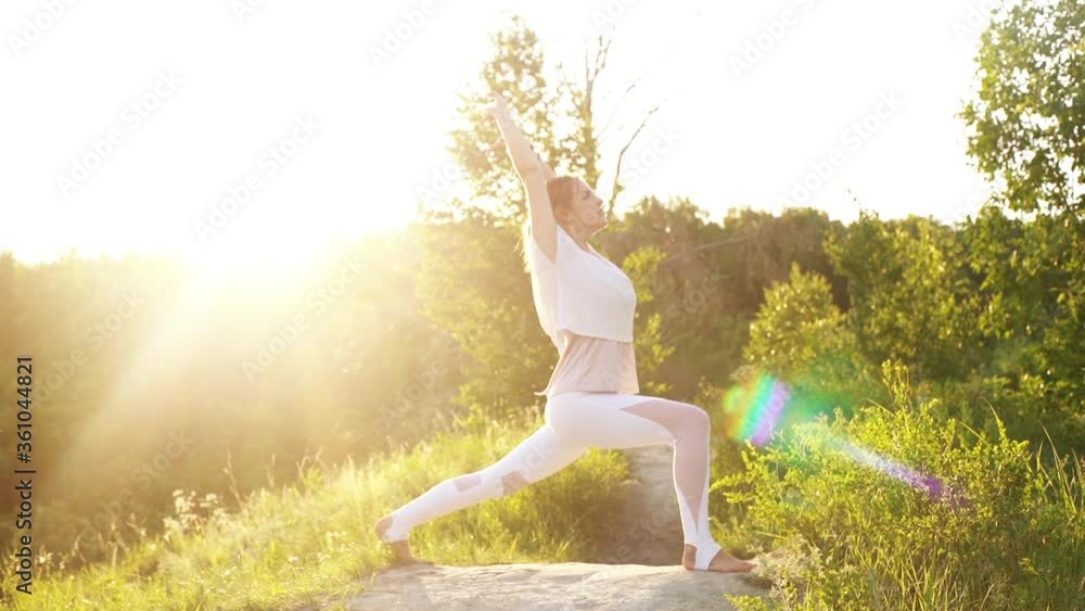 Young woman is practicing yoga and performing Warrior Pose outside in park evening on background of sunlight. Female doing yoga exercises outdoors at green grass during sunset, slowmotion. Young woman is practicing yoga and performing Warrior Pose outside in park evening on background of sunlight. Female doing yoga exercises outdoors at green grass during sunset, slowmotion.