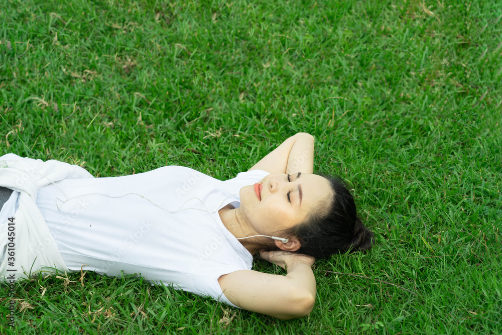 Healthy young beautiful asian woman lying and rest listening to music on earphone after running or jogging in the park in sunshine on beautiful summer day. Sport fitness training outdoor concept