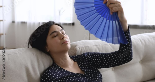 Close up view overheated asian woman lying on couch at hot summer weather day feel unwell suffers from heat wave blue fan cooling herself, dwelling without air conditioner need climate control concept