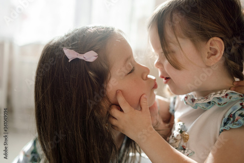 Obraz na plátně Two cute sisters of preschooler age pull their lips together to kiss each other in dreamy soft backlight