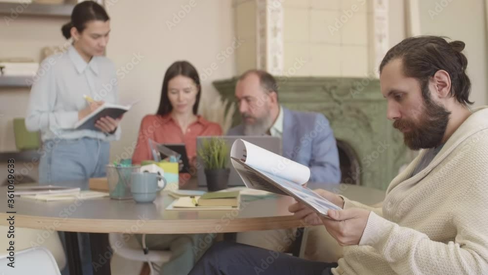 Medium shot of Caucasian bearded man wearing casual clothes sitting at desk in coworking space and looking through charts while three people working on background Medium shot of Caucasian bearded man wearing casual clothes sitting at desk in coworking space and looking through charts while three people working on background