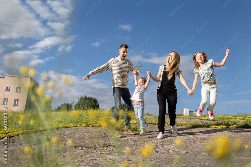 Young parents in casual look have fun and jump with their daughters in a yellow field enjoying the fresh clean air and blue sky. Blonde mother in black jumpsuit. Young parents in casual look have fun and jump with their daughters in a yellow field enjoying the fresh clean air and blue sky. Blonde mother in black jumpsuit.
