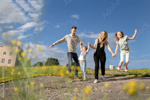 Young parents in casual look have fun and jump with their daughters in a yellow field enjoying the fresh clean air and blue sky. Blonde mother in black jumpsuit.