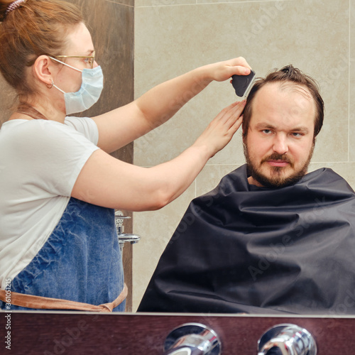 A woman in medical mask cuts a man hair with a clipper, mirror bathroom
