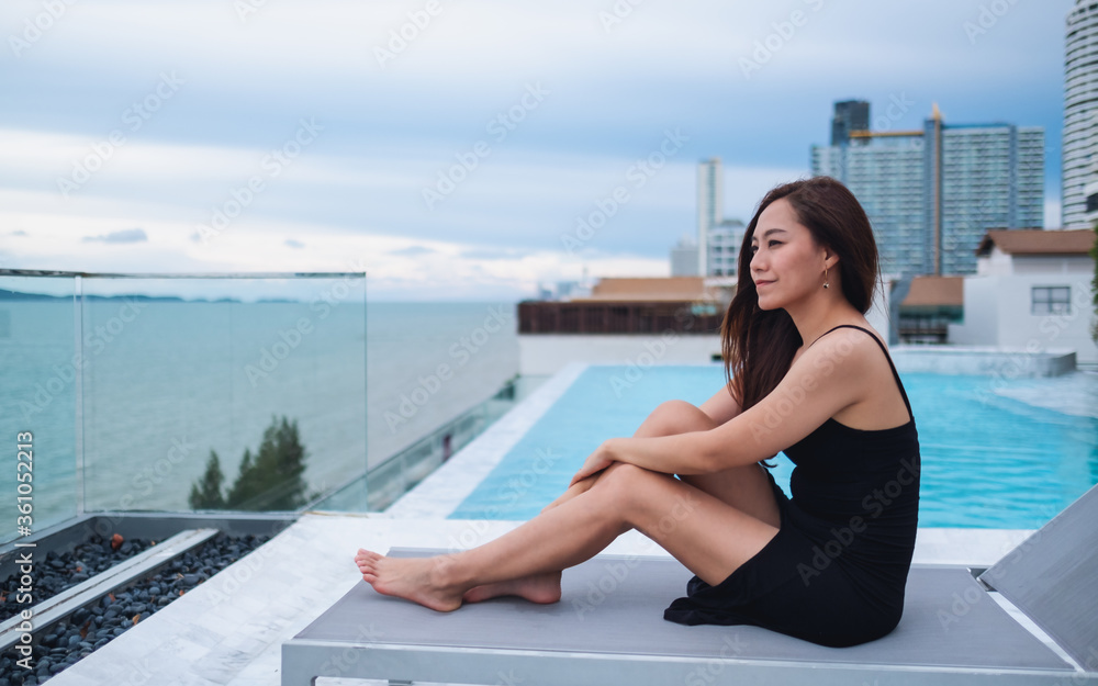 A beautiful young asian woman sitting next to the rooftop pool , looking at the sea and blue sky view