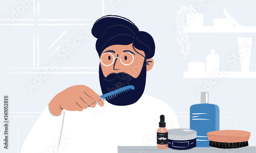 Young man wearing glasses brushing his beard and looking in the mirror having styling cosmetic, brushes and odecolone bottle in his bathroom. Everyday morning routine. Men hygiene and self care.