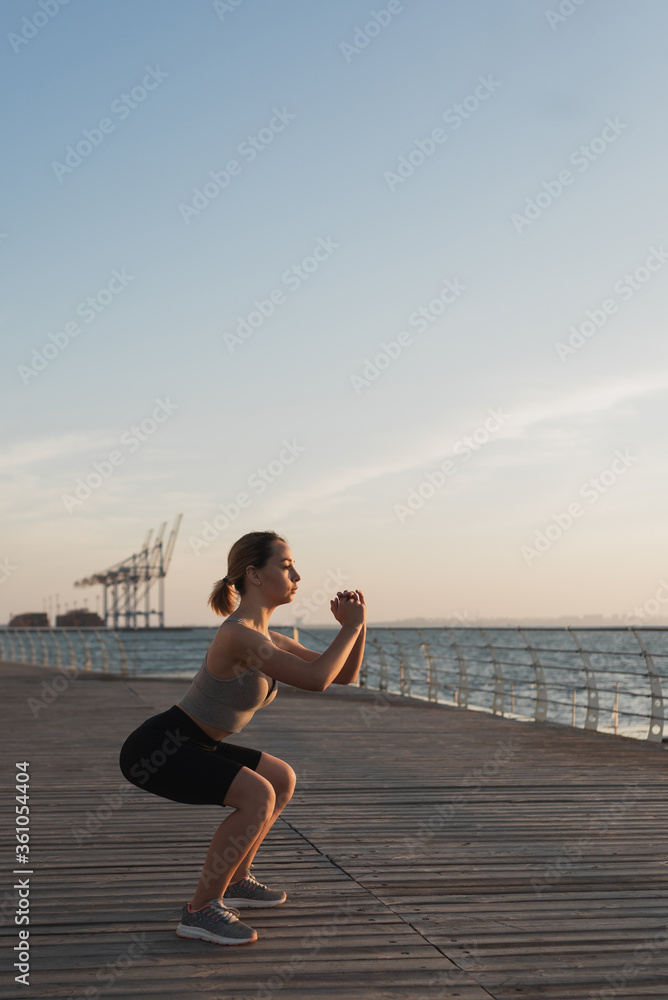 © Andrii Lysenko - the girl is engaged in fitness by the sea or ocean, crouches and smiles, in the morning light the concept of an active lifestyle.