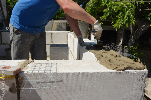A mason or building contractor is laying a brick wall from concrete autoclaved aerated blocks, applying mortar using a trowel and reinforcing wire mesh to build home addition.