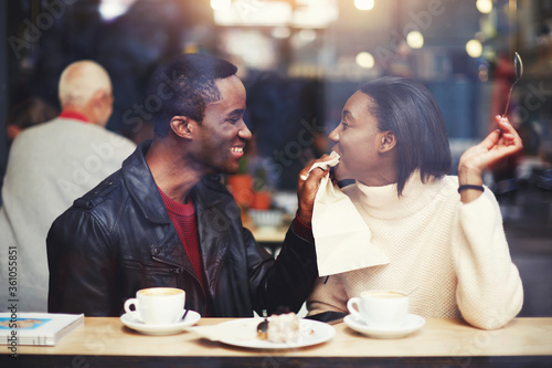 Smiling boyfriend wiping mouth with a napkin his girlfriend during breakfast in modern coffee shop interior, happy young romantic couple having fun together while sitting in cafe bar during free time