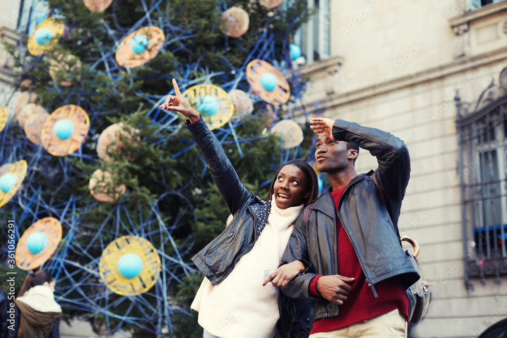 Obraz premium Charming woman pointing with he finger something interesting, romantic couple walk on christmas tree background on big square, laughing couple on christmas vacation walking in the city