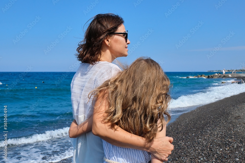 Happy mom and daughter kid hugging walking together along the beach, back view Happy mom and daughter kid hugging walking together along the beach, back view