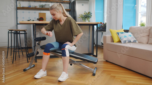 Tired caucasian sportswoman sitting on exercise bench looking on smartwatch and checking her pulse training at home. 