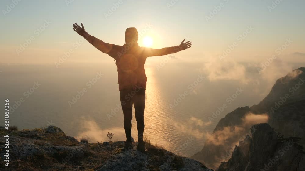 Man stands on the edge of a high mountain above the sea, spreads his arms to the sides and waves, covering his body with the shining sun, a close view. A bearded tourist stands near a cliff at sunset. Man stands on the edge of a high mountain above the sea, spreads his arms to the sides and waves, covering his body with the shining sun, a close view. A bearded tourist stands near a cliff at sunset.