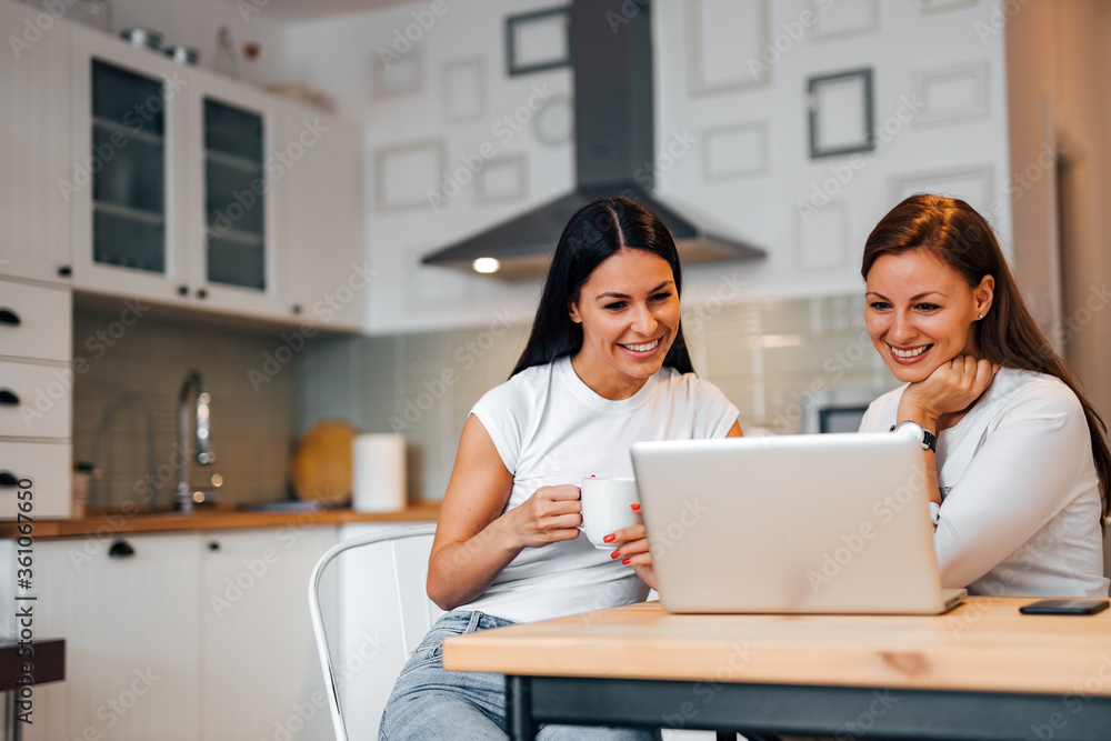 Two smiling female friends looking at laptop while sitting at kitchen table, portrait. Two smiling female friends looking at laptop while sitting at kitchen table, portrait.