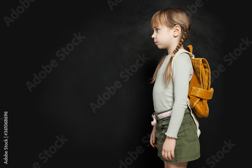 Sad school child girl standing with backpack on black. School problem concept