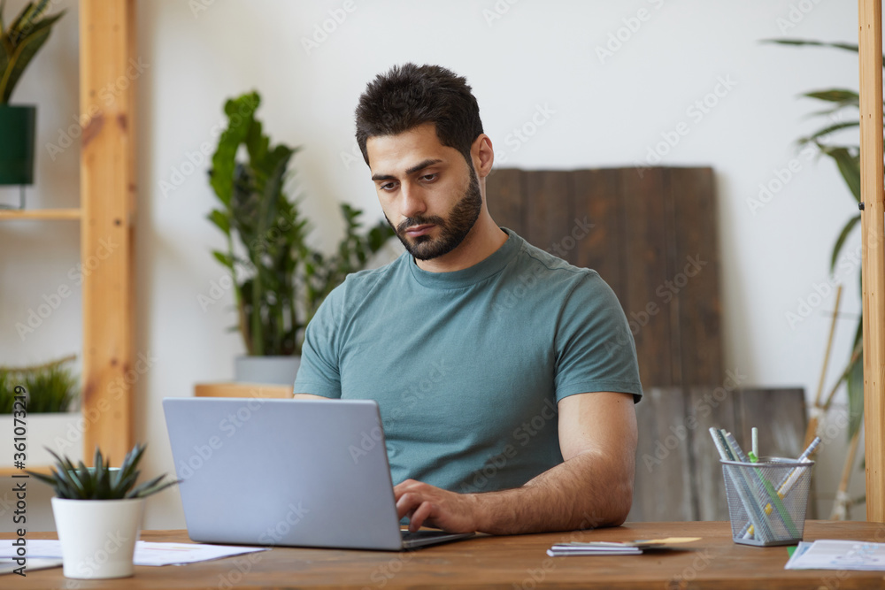 Portrait of handsome bearded man using laptop while sitting at desk in home interior decorated with houseplants, copy space