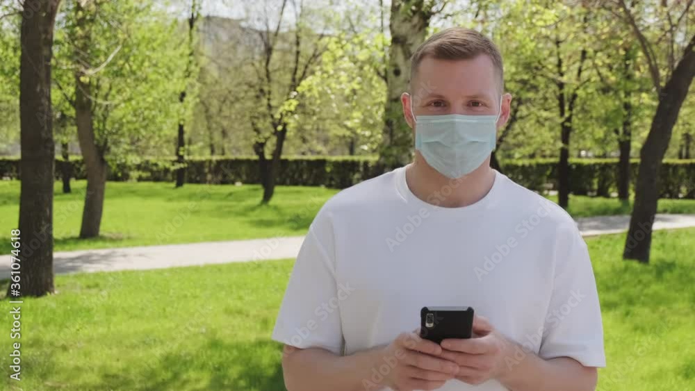 Tracking shot of young man in face mask standing outdoors in park on sunny summer day and using mobile phone, then looking at camera Tracking shot of young man in face mask standing outdoors in park on sunny summer day and using mobile phone, then looking at camera