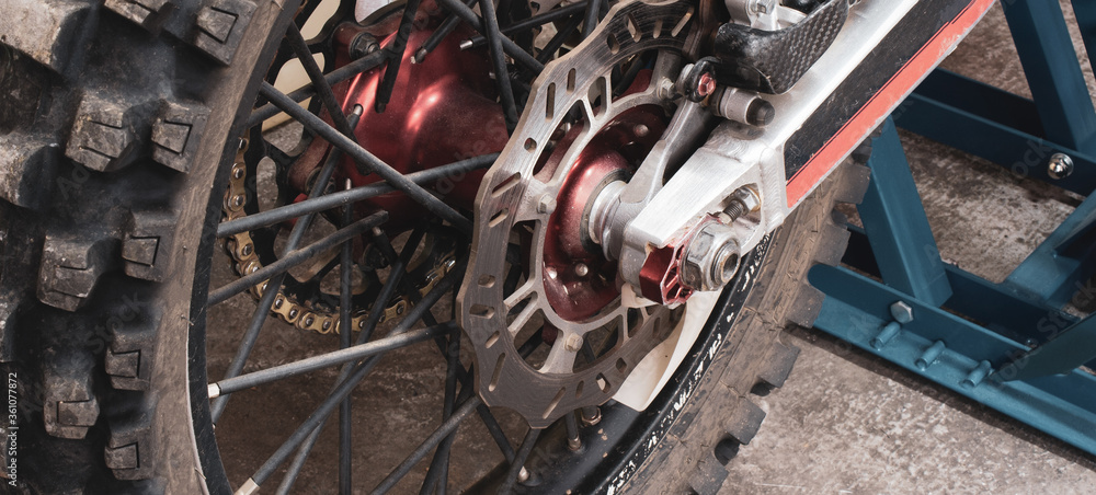 Close-up rear studded wheel of enduro off-road motorcycle in garage on lift. Brake discs, chain, transmission. Photo of a cross bike part was taken from the side in the workshop. Banner for web site Close-up rear studded wheel of enduro off-road motorcycle in garage on lift. Brake discs, chain, transmission. Photo of a cross bike part was taken from the side in the workshop. Banner for web site