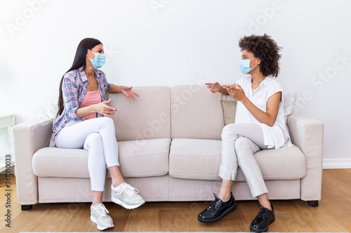Two female best friends sitting in social distance wearing face mask and talking on the sofa, preventing covid 19 coronavirus pandemic infection spread.