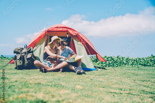 Hipster couple resting in tent camping on green grass during sunny summer day planning route,young woman pointing on map discussing direction for exploring nature with boyfriend on vacations