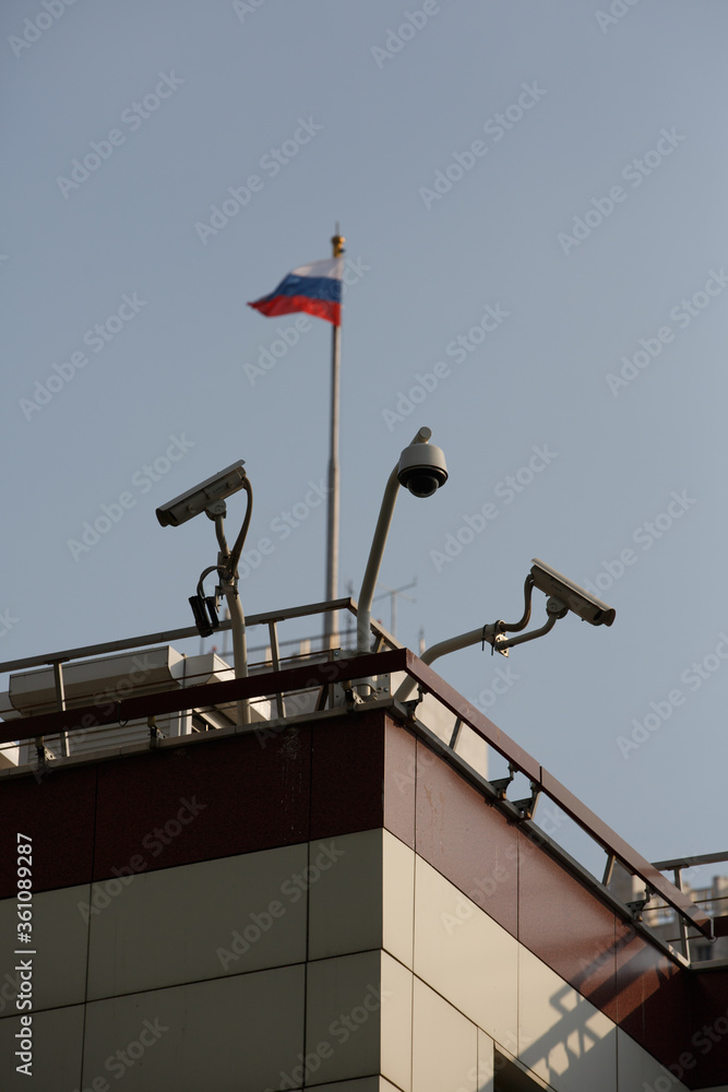 vertical day shot of three cctv surveillance cameras on the walls of the Parlament and a flag of Russia on a blue sky background vertical day shot of three cctv surveillance cameras on the walls of the Parlament and a flag of Russia on a blue sky background