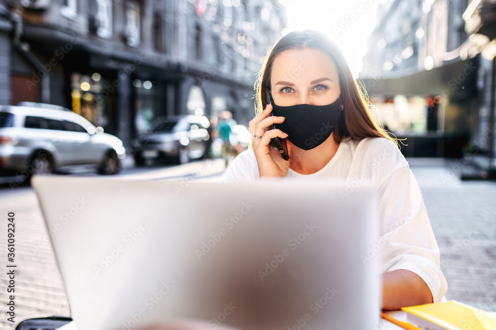 Obraz premium Close-up portrait of young woman with a protective mask at the table on the face on the summer terrace. Young woman works remotely with laptop in cafe, she looks at camera and talks on the smartphone