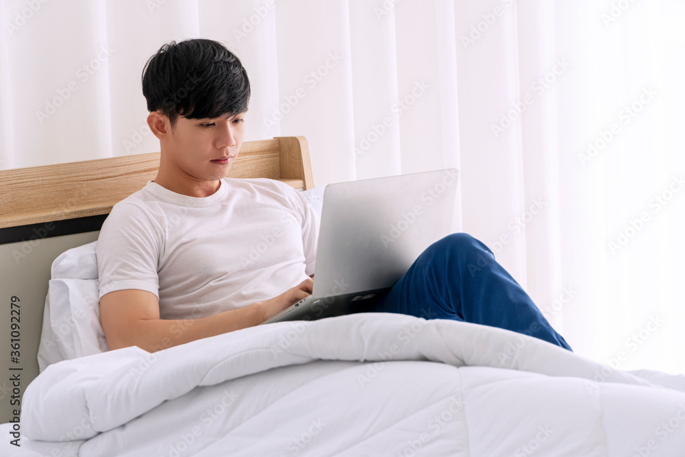 Freelance young man using laptop working on internet while sitting on the white bed with pillow in the morning at apartment. Lifestyle work at home with technology concept. Freelance young man using laptop working on internet while sitting on the white bed with pillow in the morning at apartment. Lifestyle work at home with technology concept.