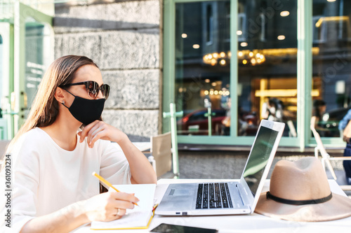 Female student is watching lectures on laptop and taking notes while sitting at the table with a medical mask on the face in the summer street cafe