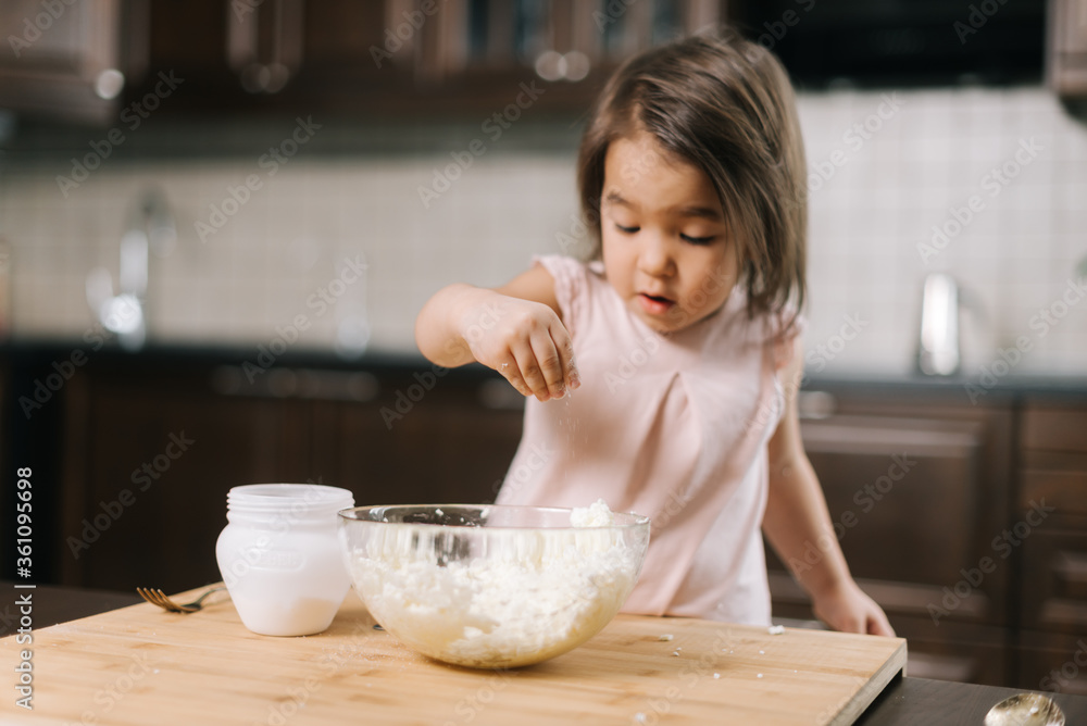 Playful beautiful little girl is taking salt from the salt cellar with fingers to add to the dough in transparent bowl for making cheesecakes at the table in kitchen with modern interior.