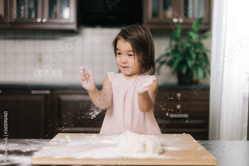 Emotional beautiful little girl is clapping hands with flour. Child is playing with flour in the kitchen while cooking homemade baked goods.
