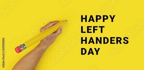 фотографія Big pencil in male left hand on yellow background with text Happy Left Handers Day