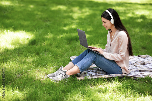 Smiling asian girl relaxing with laptop and headphones on lawn in park