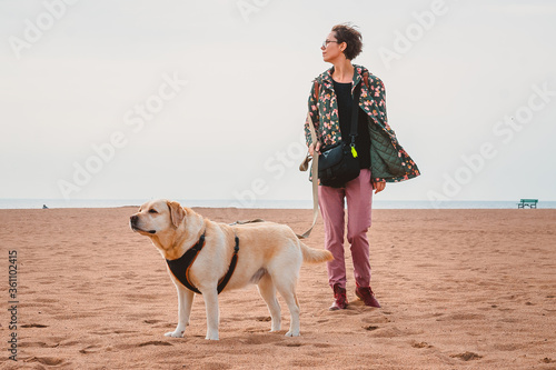A girl and a fawn Labrador Retriever walk on a sandy beach. Traveling with a dog on the sea