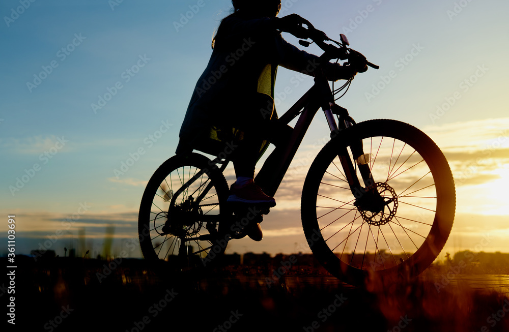 Obraz premium Girl is cycling road bike in the sunset A young girl is cycling into the sunset in the park Silhouette of a teenager rides a bike at sunset. Orange-blue sky background.