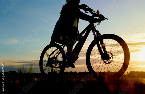 Fototapeta Naklejka Na Ścianę i Meble -  Girl is cycling road bike in the sunset A young girl is cycling into the sunset in the park Silhouette of a teenager rides a bike at sunset. Orange-blue sky background.