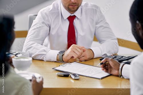 black clients sign a contract while sitting at table with salesman, professional worker of dealership who is waiting them
