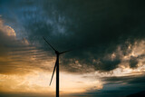 The silhouette of a high industrial wind turbine against the sunset sky with clouds in the evening.