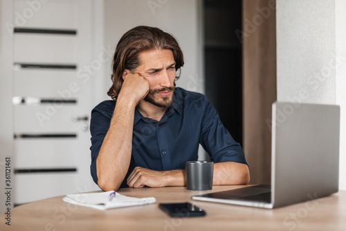 Pensive young man thinking about solving a problem stuck with a task while working with a laptop remotely. A man works at home does not know how to solve a problem